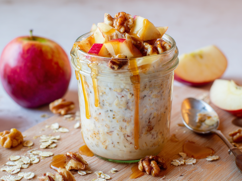 Ein Glas Apfelkuchen Overnight Oats steht auf einem Holzbrett. Es ist mit Apfelstücken, Walnüssen und Ahornsirup getoppt. Im Hintergrund liegen mehrere Äpfel, die meisten davon kleingeschnitten. Um das Glas verteilt liegen Haferflocken und Walnüsse.