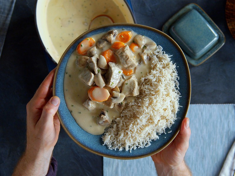 Ein dunkelblauer Teller mit einer Portion Blanquette de Veau mit Reis. Der Teller wir von zwei Händen gehalten. Draufsicht. Unter dem Teller kann man den Kochtopf mit dem Blanquette de Veau erkennen, sowie einen hellblauen Tischläufer, Salz und Pfefferstreuer und Besteck sowie sonstige Küchenutensilien.