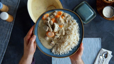 Ein dunkelblauer Teller mit einer Portion Blanquette de Veau mit Reis. Der Teller wir von zwei Händen gehalten. Draufsicht. Unter dem Teller kann man den Kochtopf mit dem Blanquette de Veau erkennen, sowie einen hellblauen Tischläufer, Salz und Pfefferstreuer und Besteck sowie sonstige Küchenutensilien.