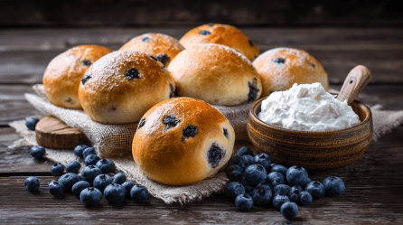 Ein Brett mit sieben Heidelbeer-Quarkbrötchen. Quark und frische Heidelbeeren liegen als Deko außen herum.