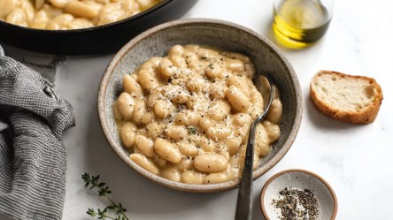 Eine Schüssel mit weißen Bohnen à la Cacio e Pepe. Ein Löffel ist in der Schüssel. Um die Schüssel herum kann man eine kleine Schale mit gemahlenem Pfeffer, ein Glasgefäß mit Olivenöl sowie eine Scheibe Brot sehen. Im Hintergrund steht eine große Pfanne mit weißen Bohnen à la Cacio e Pepe.