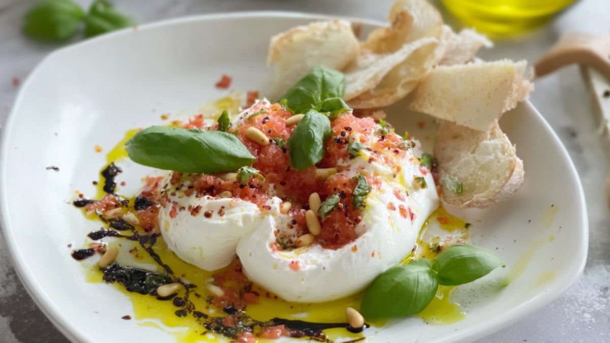 Eine Portion Burrata mit gefrorener Tomate. Das Ganze ist mit Olivenöl, Balsamico, Pinienkernen und frischem Basilikum angerichtet. Im Teller liegen ein paar selbst gemachte knusprige Brotchips.