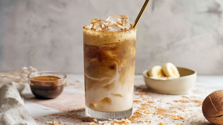 Ein großes Glas mit einem Caramelized Banana Latte. Ein goldener Strohhalm guck aus dem Glas. Im Hintergrund kann man eine Schale mit Bananenscheiben und eine mit Kaffee sehen. Außerdem liegt eine Kokosnuss im Bild. Kokosflocken und Zimt liegen verstreut herum.