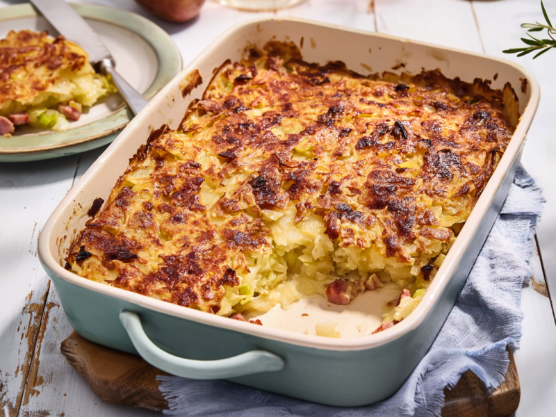 Eine Auflaufform mit einem Döppekuchen steht auf einem Brett auf einem blauen Tuch. Links im Hintergrund ist ein Teller mit einem Stück des Auflaufs. Rechts im Bild sieht man frischen Rosmarin.