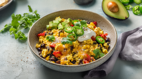 Eine Schale mit Enchilada-Rührei steht auf einem grauen Tisch. Darum verteilt liegen einige Zutaten und rechts neben der Schale ein graues Tuch.