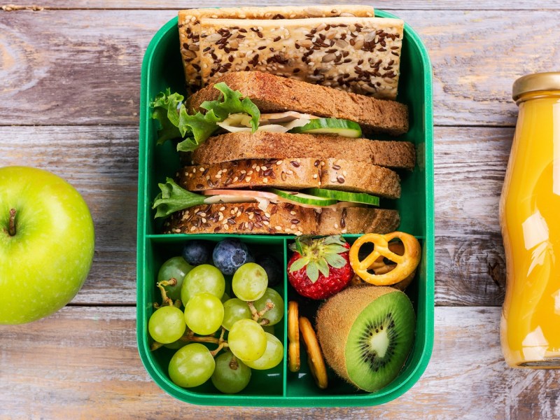 7 Tipps für Essen auf Reisen: Auf einem holzfarbenen Untergrund sind eine Lunchbox, ein Apfel und eine Flasche zu sehen.