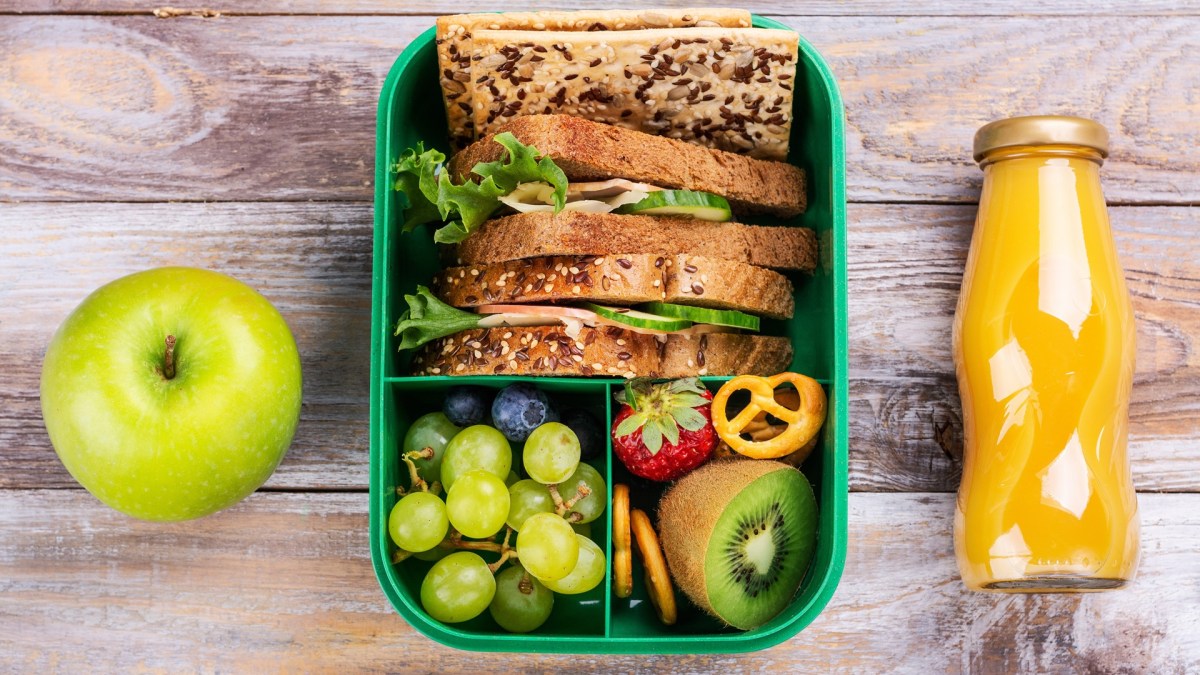 7 Tipps für Essen auf Reisen: Auf einem holzfarbenen Untergrund sind eine Lunchbox, ein Apfel und eine Flasche zu sehen.