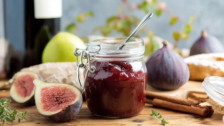 Ein Einmachglas mit Feigen-Rotwein-Marmelade, in dem ein Löffel steckt. Ringsum liegen halbierte und ganze Feigen, Zimtstangen, Thymian und eine Zitrone. Im Hintergrund kann man eine Rotweinflasche erkennen.