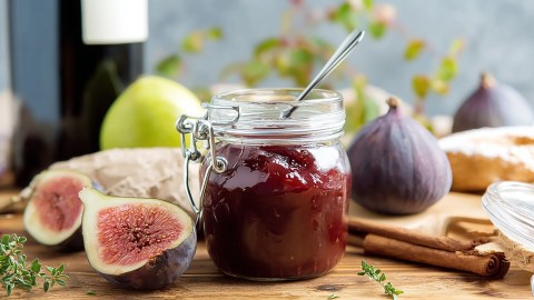 Ein Einmachglas mit Feigen-Rotwein-Marmelade, in dem ein Löffel steckt. Ringsum liegen halbierte und ganze Feigen, Zimtstangen, Thymian und eine Zitrone. Im Hintergrund kann man eine Rotweinflasche erkennen.