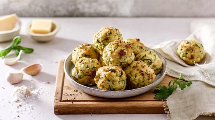 Ein Teller, der auf einem Holzbrett steht und in dem einige gebackene Zucchini-Knödel liegen. Außerdem kann man im Bild zwei kleine Schalen mit jeweils einem Stück Butter, Knoblauchzehen und verstreutes Mehl sehen.