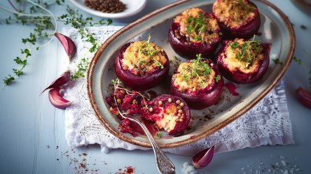 Eine Auflaufform mit einigen gefüllten Roten Beten. Die Form steht auf einem Stoffuntersetzer. Ringsum liegen einige Knoblauchzehen sowie frische Kräuter.