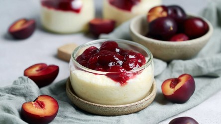 Eine kleine Glasschüssel mit Grießquark mit Pflaumenkompott, die auf einem Untersetzer steht. Ringsum liegen halbierte und entsteinte Pflaumen und einige Schalen mit Pflaumen sind zu sehen.