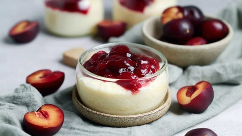 Eine kleine Glasschüssel mit Grießquark mit Pflaumenkompott, die auf einem Untersetzer steht. Ringsum liegen halbierte und entsteinte Pflaumen und einige Schalen mit Pflaumen sind zu sehen.