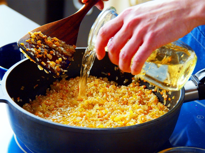 Grundrezept für Risotto: Eine Köchin gießt Weißwein zu Risottoreis.