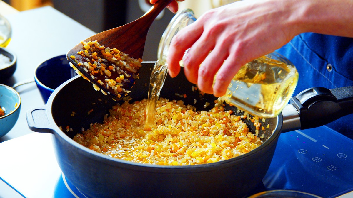 Grundrezept für Risotto: Eine Köchin gießt Weißwein zu Risottoreis.