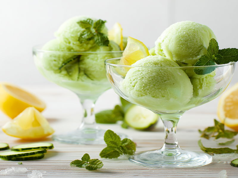 Zwei Dessertgläser mit Gurken-Minz-Sorbet. Um die Gläser verteilt liegen einige Gurkenscheiben, Zitronenstücke und Minzblätter.