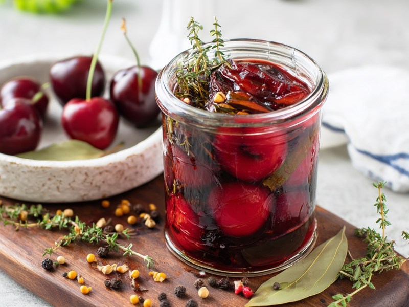 Ein Glas mit herzhaft eingelegten Kirschen. Drumherum liegen Pfeffer- und Senfkörner sowie frischer Thymian und Lorbeerblätter. Im Hintergrund ist eine Schale mit frischen Kirschen erkennbar.