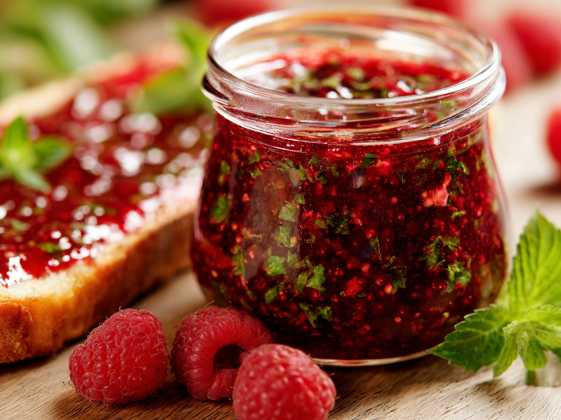 Ein Glas mit Himbeer-Minz-Marmelade auf einem Tisch. Davor liegen drei Himbeeren, rechts daneben ein Minzblatt, links daneben ein Brot mit der Marmelade.