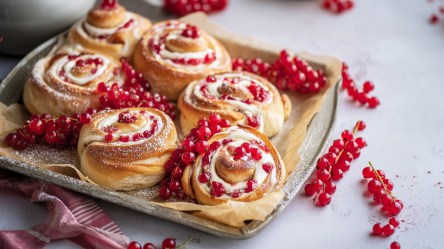 Ein kleines Backblech mit Johannisbeerschnecken. Das Ganze ist mit frischen roten Johannisbeeren garniert und dekoriert. Auch neben dem Blech liegen einige Beeren verteilt.