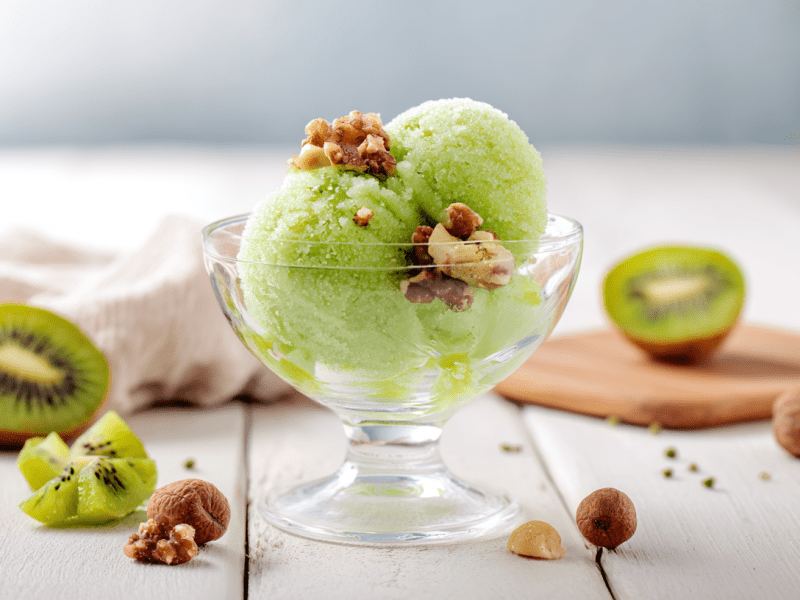 Ein Glas Zitronensorbet auf einem weißen Holztisch. Um das Glas herum liegen einige Nüsse verteilt. Im Hintergrund liegen zwei Kiwihälften.