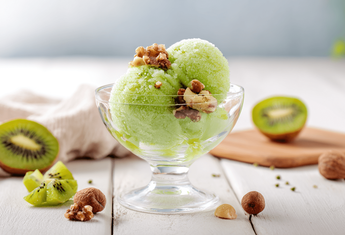 Ein Glas Zitronensorbet auf einem weißen Holztisch. Um das Glas herum liegen einige Nüsse verteilt. Im Hintergrund liegen zwei Kiwihälften.