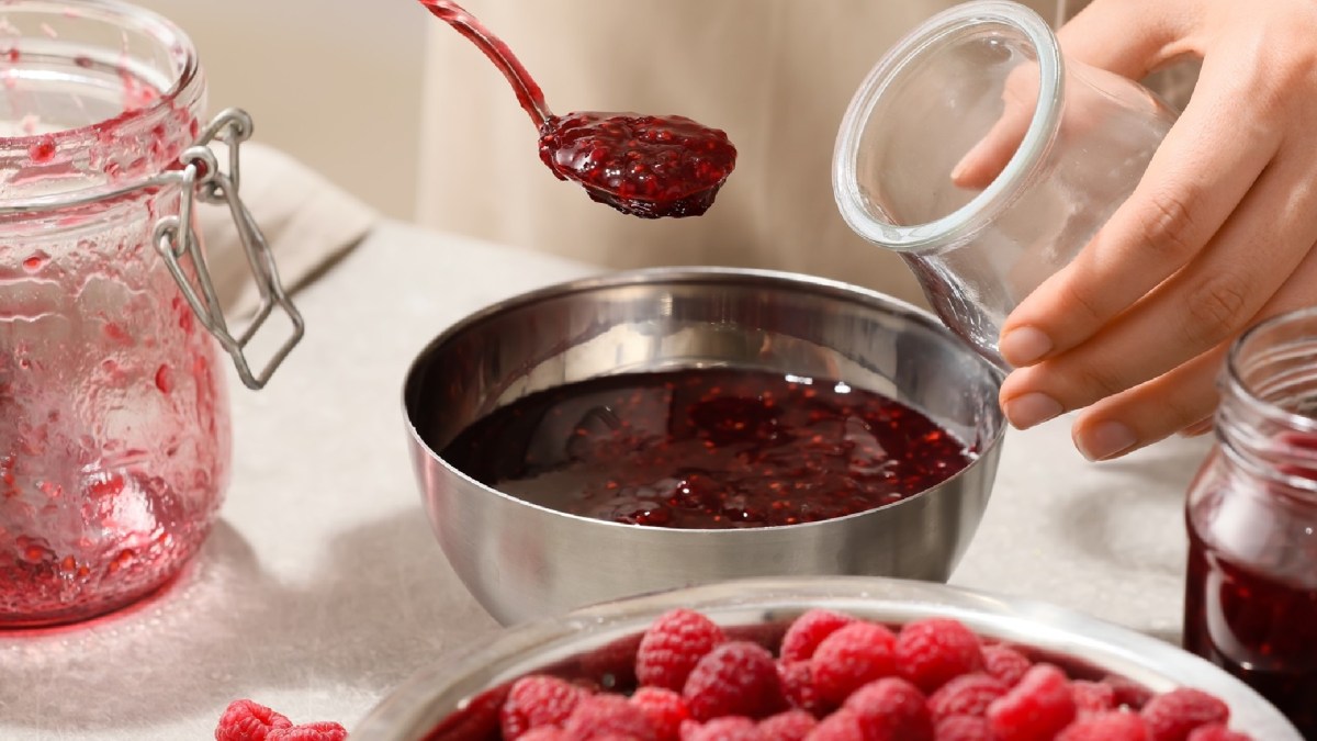 Marmeladen-Zubehör: Ein Mensch füllt Himbeermarmelade aus einer silbernen Schale in ein Glas. Im Vordergrund steht eine Schale mit ganzen, frischen Himbeeren.