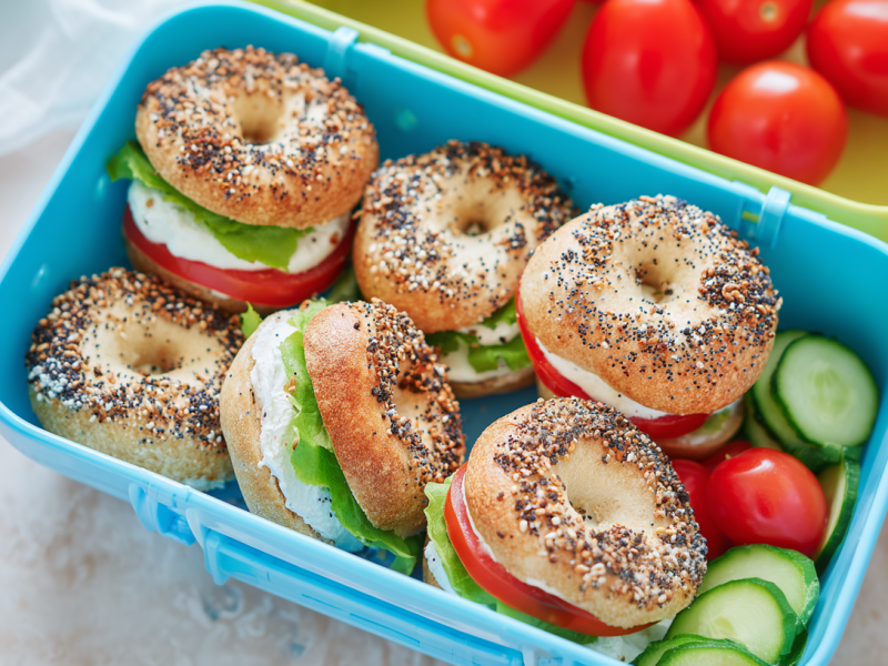 Sechs Mini-Bagel in einer blauen Brotdose. Sie sind mit Frischkäse, Tomaten und Salat belegt. In der Dose liegen noch Gurkenscheiben sowie Cherrytomaten.