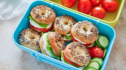 Sechs Mini-Bagel in einer blauen Brotdose. Sie sind mit Frischkäse, Tomaten und Salat belegt. In der Dose liegen noch Gurkenscheiben sowie Cherrytomaten.
