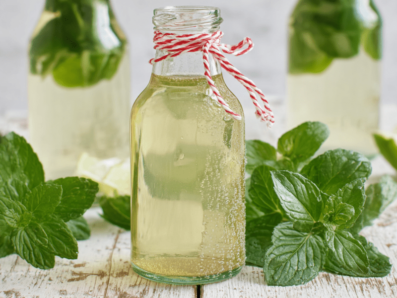 Eine Flasche mit Minz-Sirup steht im Fokus. Links und rechts daneben liegen Minzblätter. Im Hintergrund sieht man zwei weitere Flaschen mit dem Sirup sowie einige Limettenstücke.