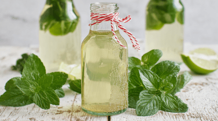 Eine Flasche mit Minz-Sirup steht im Fokus. Links und rechts daneben liegen Minzblätter. Im Hintergrund sieht man zwei weitere Flaschen mit dem Sirup sowie einige Limettenstücke.