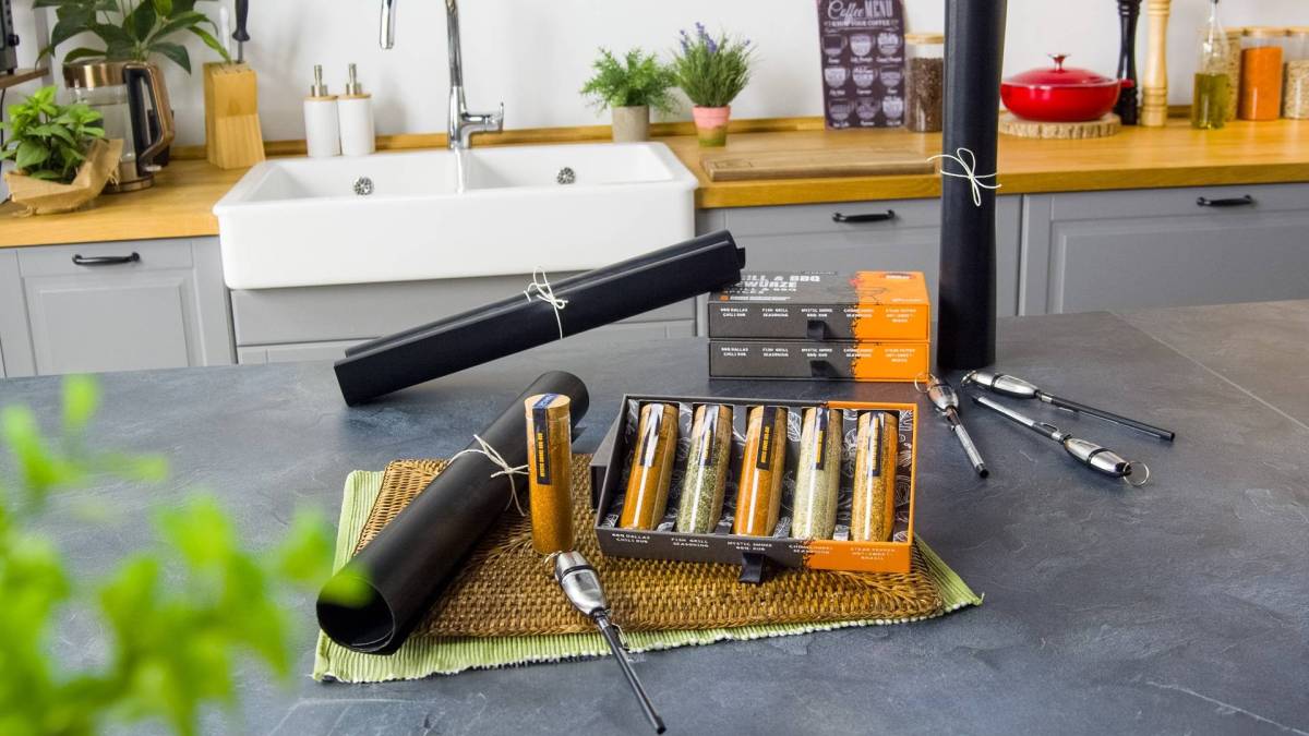 Ein Gewinnspiel-Grill-Set, das schön präsentiert wird. Es liegt eine eingerollte Grillmatte, daneben ein Fleischthermometer und eine geöffnete Schachtel mit einer 5er-Gewürmischung auf einem Tischuntersetzer. Im Vordergrund hängt verschwommen eine grüne Pflanze im Bild.