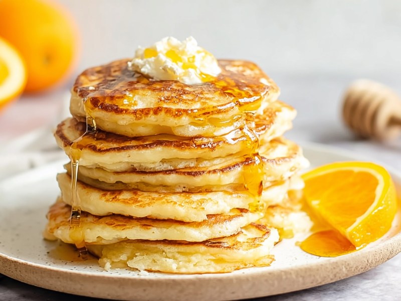 Ein Stapel mit Orangen-Ricotta-Pancakes, die mit einem kleinen Klecks Quark und Honig garniert sind. Neben den Pancakes ist eine halbe Orangenscheibe platziert. Im Hintergrund kann man eine halbe und eine ganze Orange sowie ein Honigglas und einen Honiglöffel erkennen.