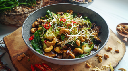 Ein Schüssel mit einer Pak Choi Pilzpfanne auf einem Holzbrett. Um die Schüssel liegen einige Zutaten wie Chilischoten und Cashewkerne verteilt.