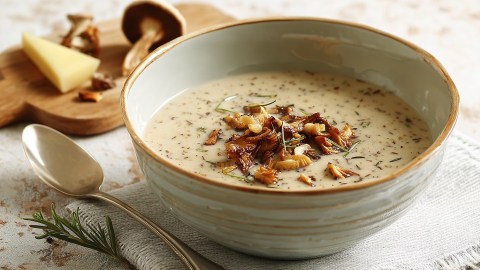 Eine Schüssel mit Parmesan-Pilzsuppe, die mit einigen gebratenen Pilzen garniert ist. Neben der Schüssel liegt ein Zweig Rosmarin und ein Löffel. Im Hintergrund sind einige Pilze auf einem kleinen Holzbrett zu sehen.