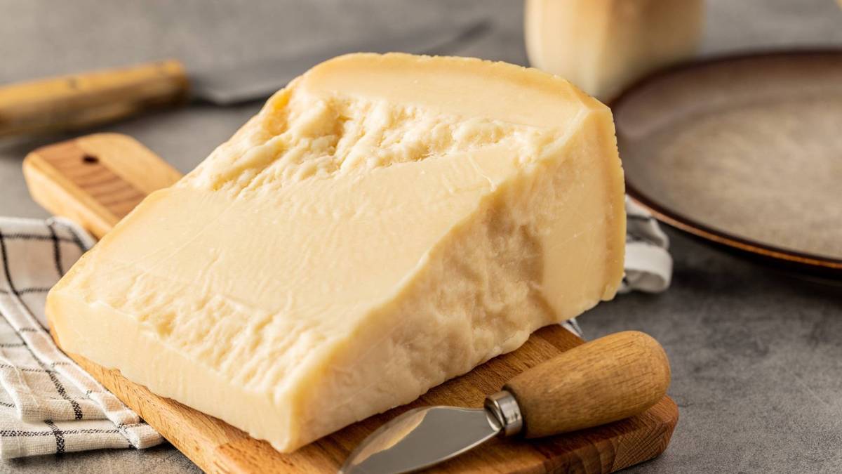 Ein Stück Hartkäse, das auf einem Holzbrett liegt. Ein Käsemesser liegt dabei. Im Hintergrund kann man einen Teller erkennen.