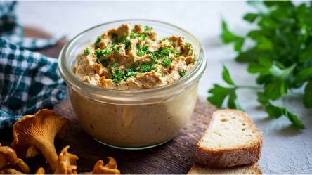 Glas mit Pfifferling-Frischkäse-Aufstrich auf einem Tisch, frische Pfifferlinge und eine Scheibe Brot daneben