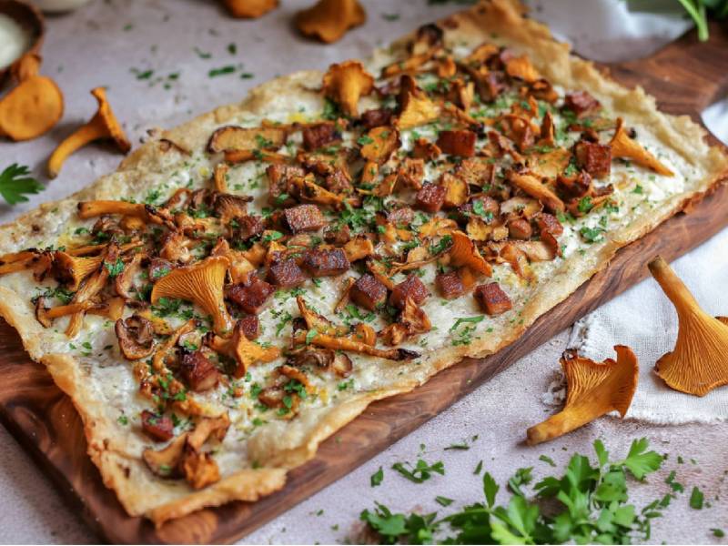 Holzbrett mit einem Pfifferling-Flammkuchen mit Speck, frische Pfifferlingen daneben