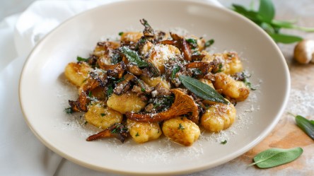 Ein großer Teller mit Pfifferling-Salbei-Gnocchi, die mit etwas frisch geriebenem Parmesan garniert sind. Links vom Teller liegt eine weiße Tischdecke drapiert. Rechts vom Teller kann man auf dem Holztisch einige frische Salbeiblätter herumliegen sehen.