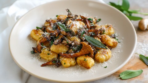 Ein großer Teller mit Pfifferling-Salbei-Gnocchi, die mit etwas frisch geriebenem Parmesan garniert sind. Links vom Teller liegt eine weiße Tischdecke drapiert. Rechts vom Teller kann man auf dem Holztisch einige frische Salbeiblätter herumliegen sehen.