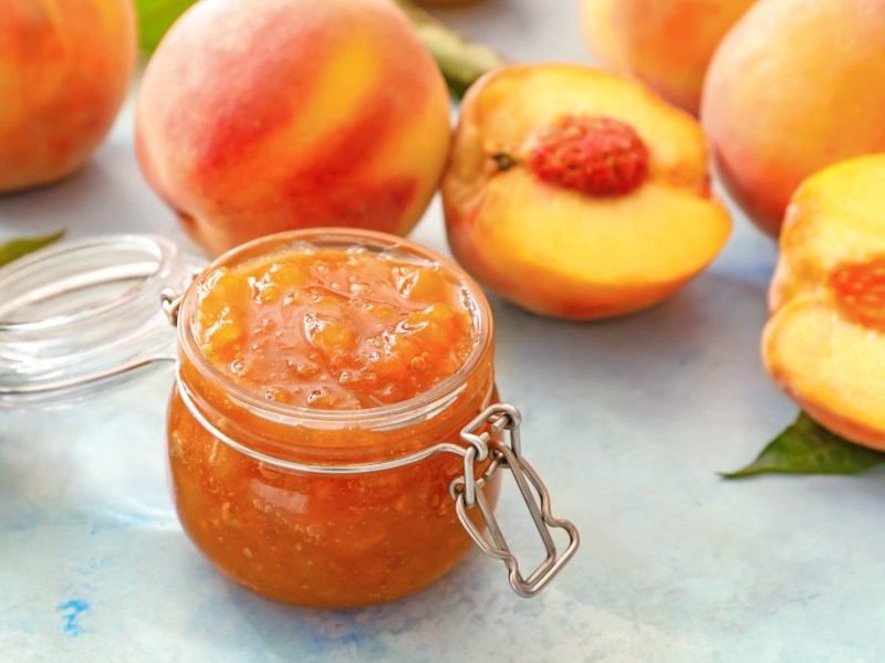 Ein Glas Pfirsichmarmelade mit Tonkabohnen. Daneben liegen frische, halbierte Pfirsiche.