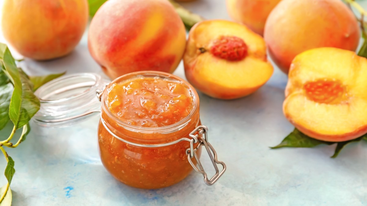 Ein Glas Pfirsichmarmelade mit Tonkabohnen. Daneben liegen frische, halbierte Pfirsiche.