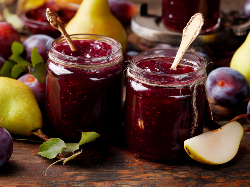 Zwei Gläser mit Pflaumen-Birnen-Marmelade. In den Gläsern stecken Löffel, außen herum liegt frisches Obst.