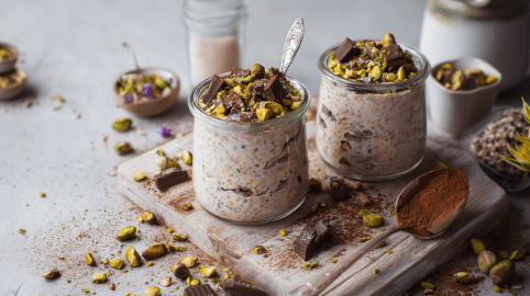 Zwei Gläser Pistazien-Schoko-Overnight Oats stehen auf einem kleinen Holzbrett. Sie haben gehackte Pistazien und Schokolade als Topping. Um sie herum liegen einige Pistazien und Schokostücke verteilt. Auf dem Brett liegt außerdem noch ein Löffel mit Kakaopulver.
