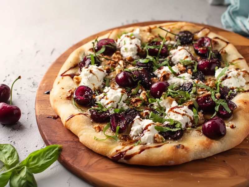 Eine Pizza mit Kirschen und Feta, die auf einem runden Holzbrett liegt. Neben dem Brett liegen einige frische Basilikumblätter und frische Kirschen.