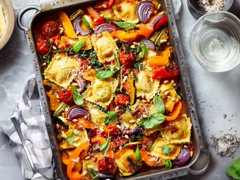 Ravioli mit Gemüse vom Blech. Das Blech steht auf einer hellen Arbeitsfläche teils af einem grauen Geschirrtuch- Ringsum liegt frisches Basilikum, einige Tomaten. Es steht ein Glas Wasser da und eine Schale mit großen dünnen Parmesanscheiben.