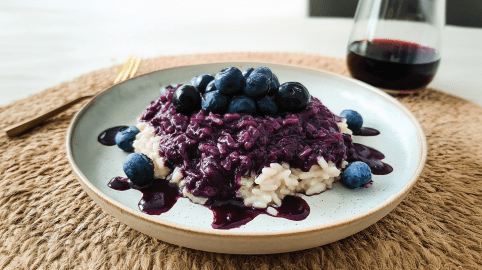 Ein Teller mit Risotto mit Heidelbeeren. Daneben liegt eine Gabel, dahinter steht ein Glas Rotwein.