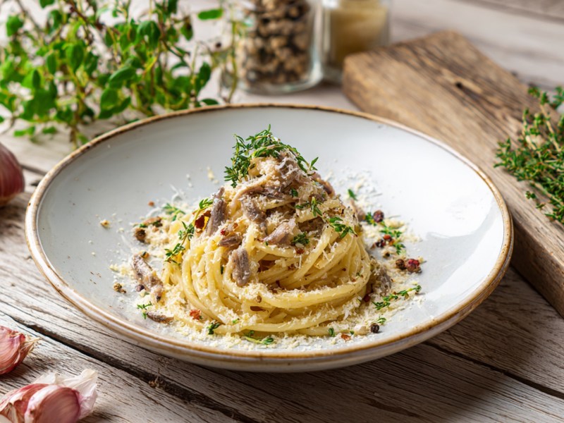 Ein Teller mit Sardellen-Carbonara, die mit frischen Kräutern und Parmesan garniert ist. Ringsum liegen eine Knoblauchknolle, zwei Knoblauchzehen, frische Kräuter herum. Auch eine Pfeffermühle ist halb erkennbar.