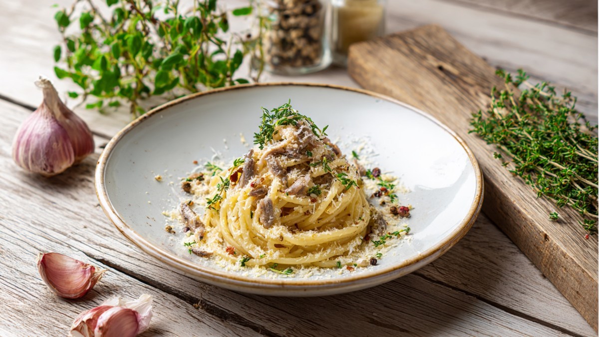 Ein Teller mit Sardellen-Carbonara, die mit frischen Kräutern und Parmesan garniert ist. Ringsum liegen eine Knoblauchknolle, zwei Knoblauchzehen, frische Kräuter herum. Auch eine Pfeffermühle ist halb erkennbar.