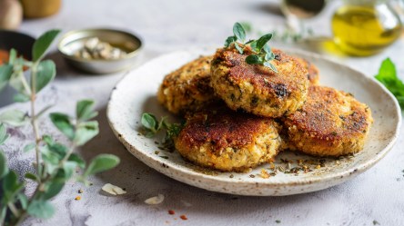 Ein Teller mit einigen Sardinen-Küchlein, die mit frischem Oregano garniert sind. Im Hintergrund kann man ein Gefäß mit Olivenöl und eine Schale mit Sardinen erkennen.