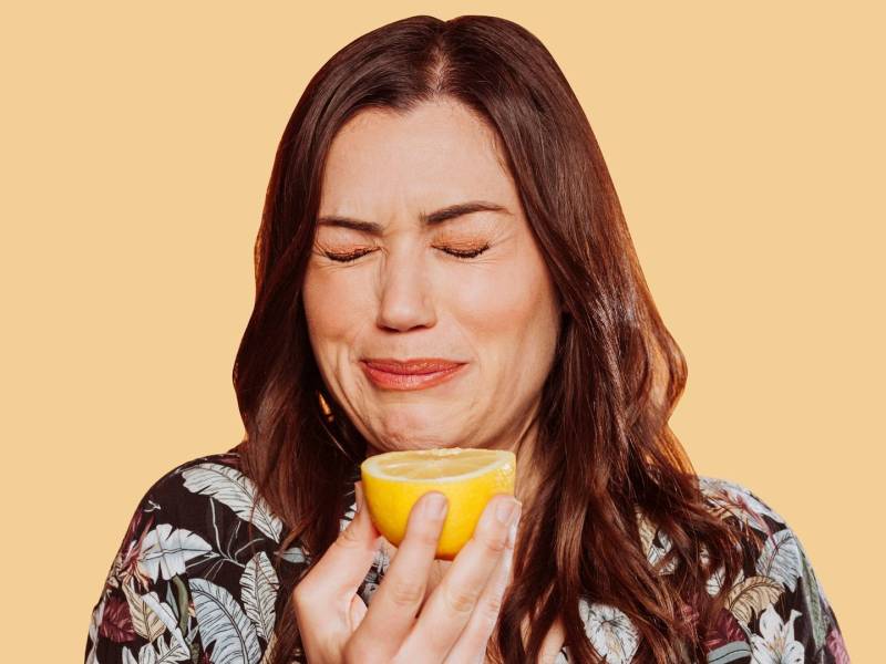 Eine Frau, die eine Grimasse mit geschlossenen Augen zieht, weil sie gerade etwas von einer Zitrone gekostet hat. Die Zitronenhälfte hält sie vor sich. Der Hintergrund ist hellorange.